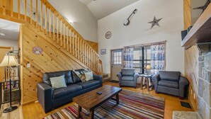 A cozy cabin interior with a wooden staircase, a blue leather sofa, two armchairs, a wooden coffee table, and a striped rug. - Divine Escape - Two Bedroom Chalet (Gatlinburg)