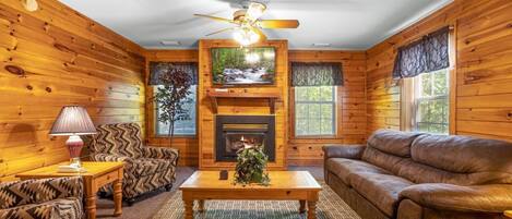 A cozy cabin interior with wooden walls, a fireplace, a ceiling fan, and a large window.