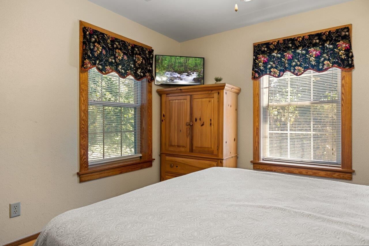 A bedroom with a bed, two windows with blinds, a wooden cabinet, and a television on the wall.