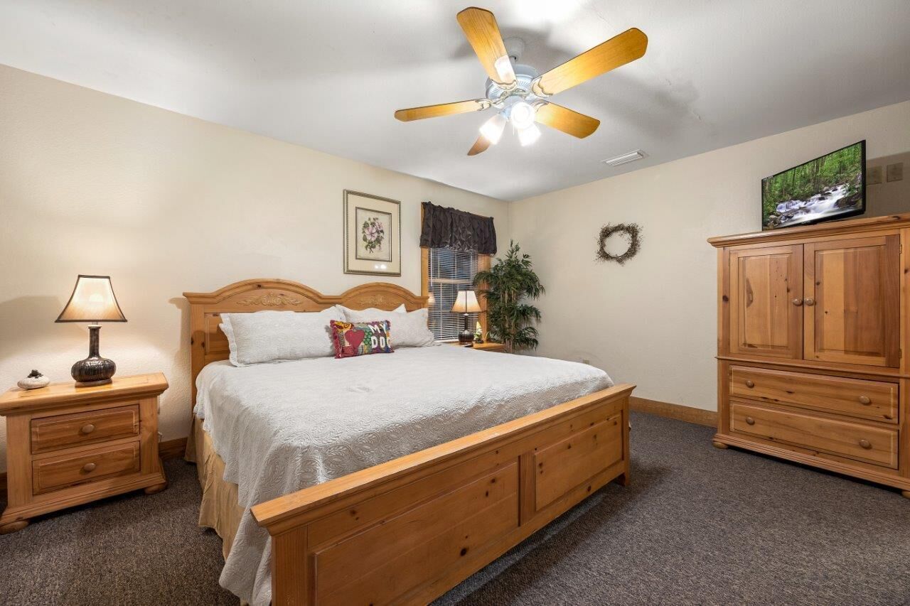 A bedroom with a wooden bed, a ceiling fan, a nightstand with a lamp, a dresser, and a television on the wall.