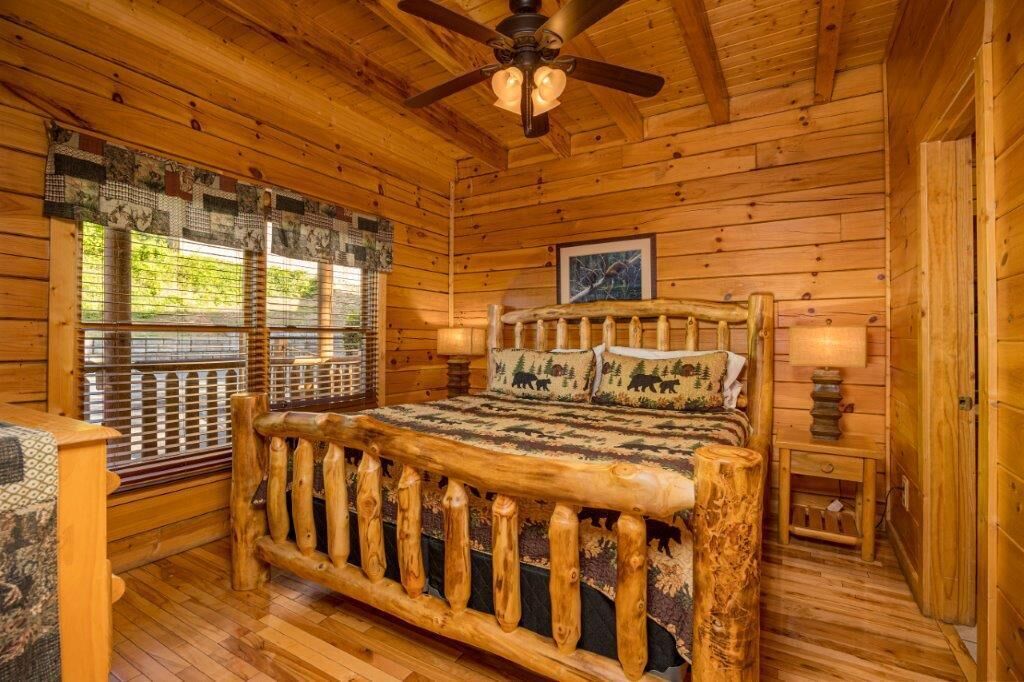 Quarto de cabana de madeira com cama, ventilador de teto e janela com vista para o verde.