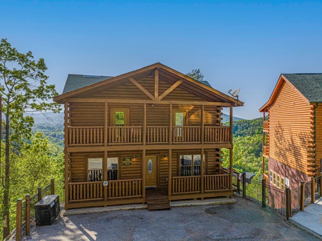 A two-story log cabin with a balcony, a front entrance, and a view of the surrounding forest.