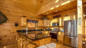 Cocina tipo cabaña de madera con una isla central, taburetes de bar, electrodomésticos de acero inoxidable y un cartel que dice 'Bienvenido'.