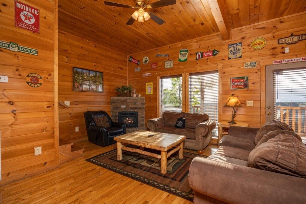 A cozy cabin interior with wooden walls, a fireplace, a ceiling fan, and various vintage signs.