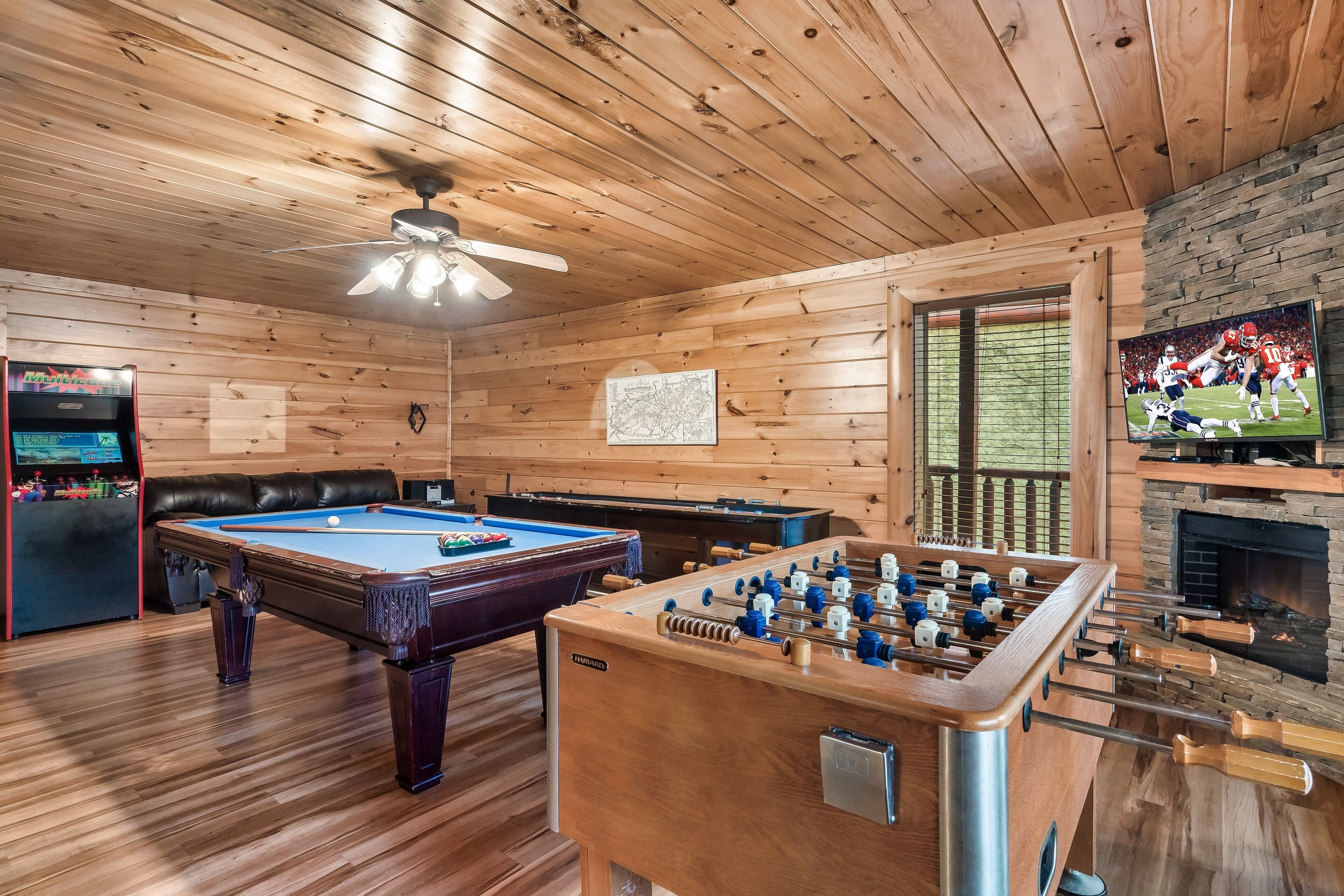 A wooden-floored room with a pool table, foosball table, and a television displaying a football game.