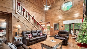 A cozy wooden cabin interior with a leather sofa, a wooden coffee table, and a staircase with red ribbons. - Mayberry - Three Bedroom Cabin (Sevierville)
