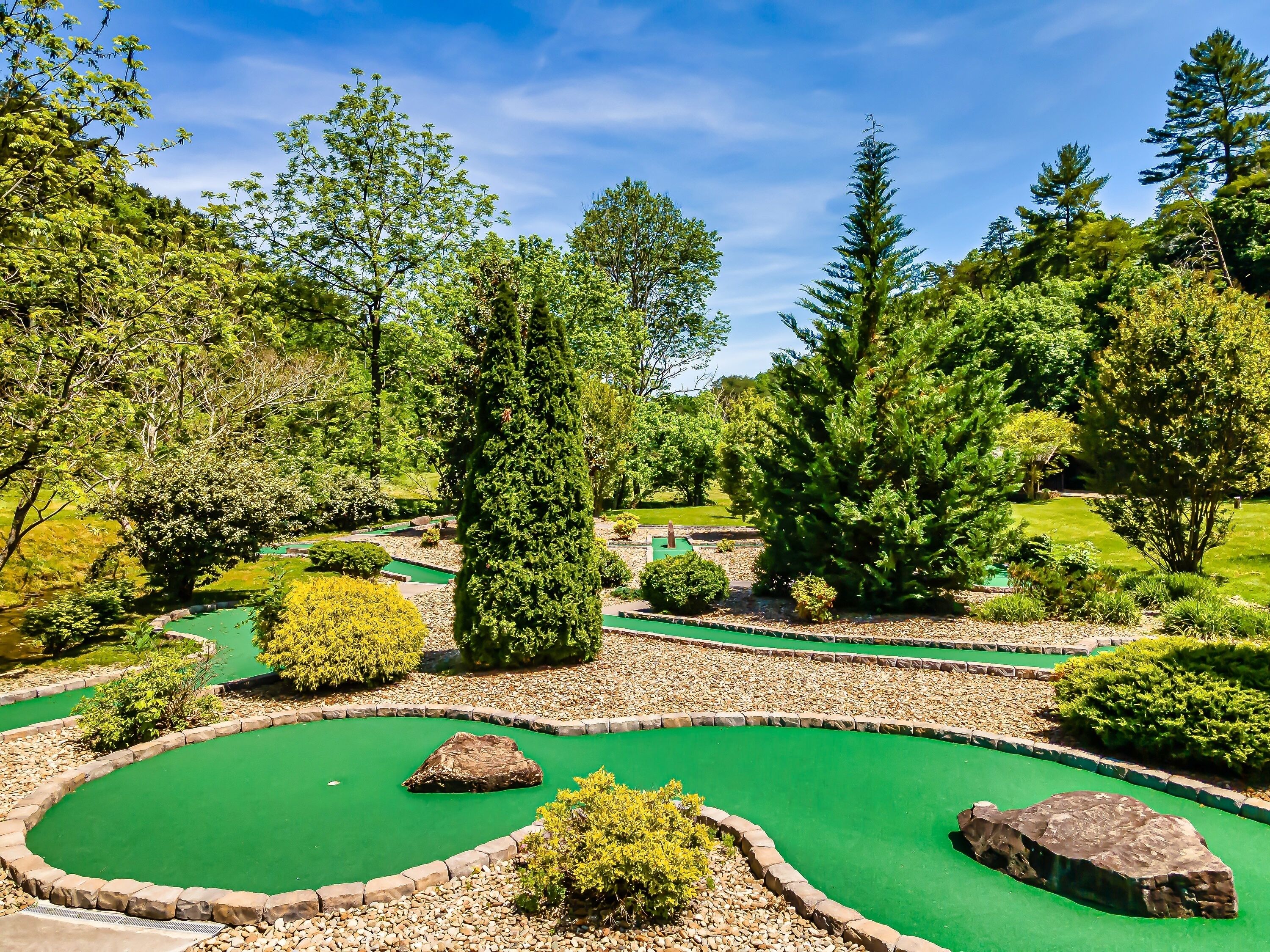 A miniature golf course with multiple green putting areas, surrounded by various trees and shrubs.