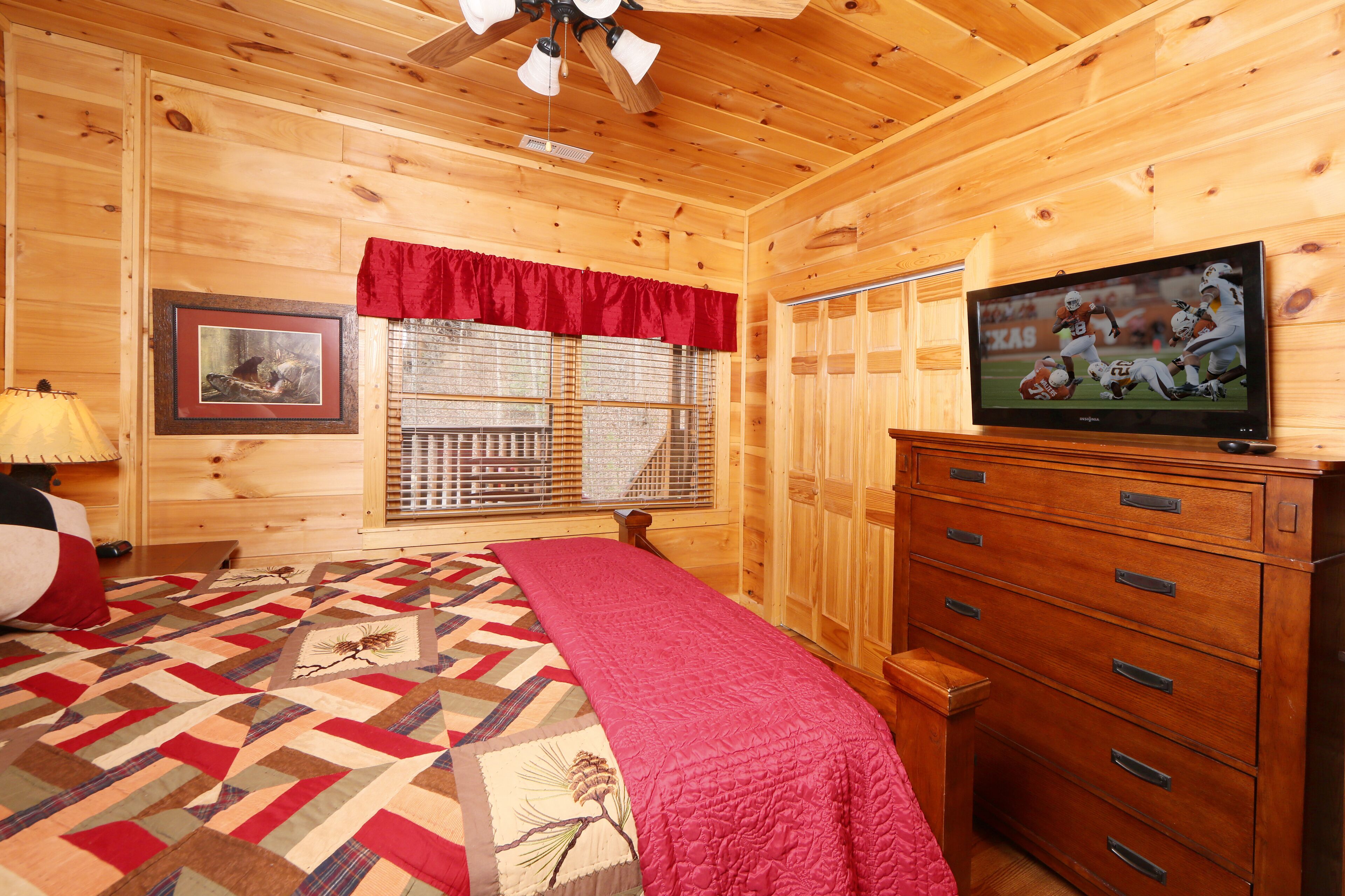 A wooden cabin room with a bed, a dresser, a television, a window with blinds, and a framed picture on the wall.