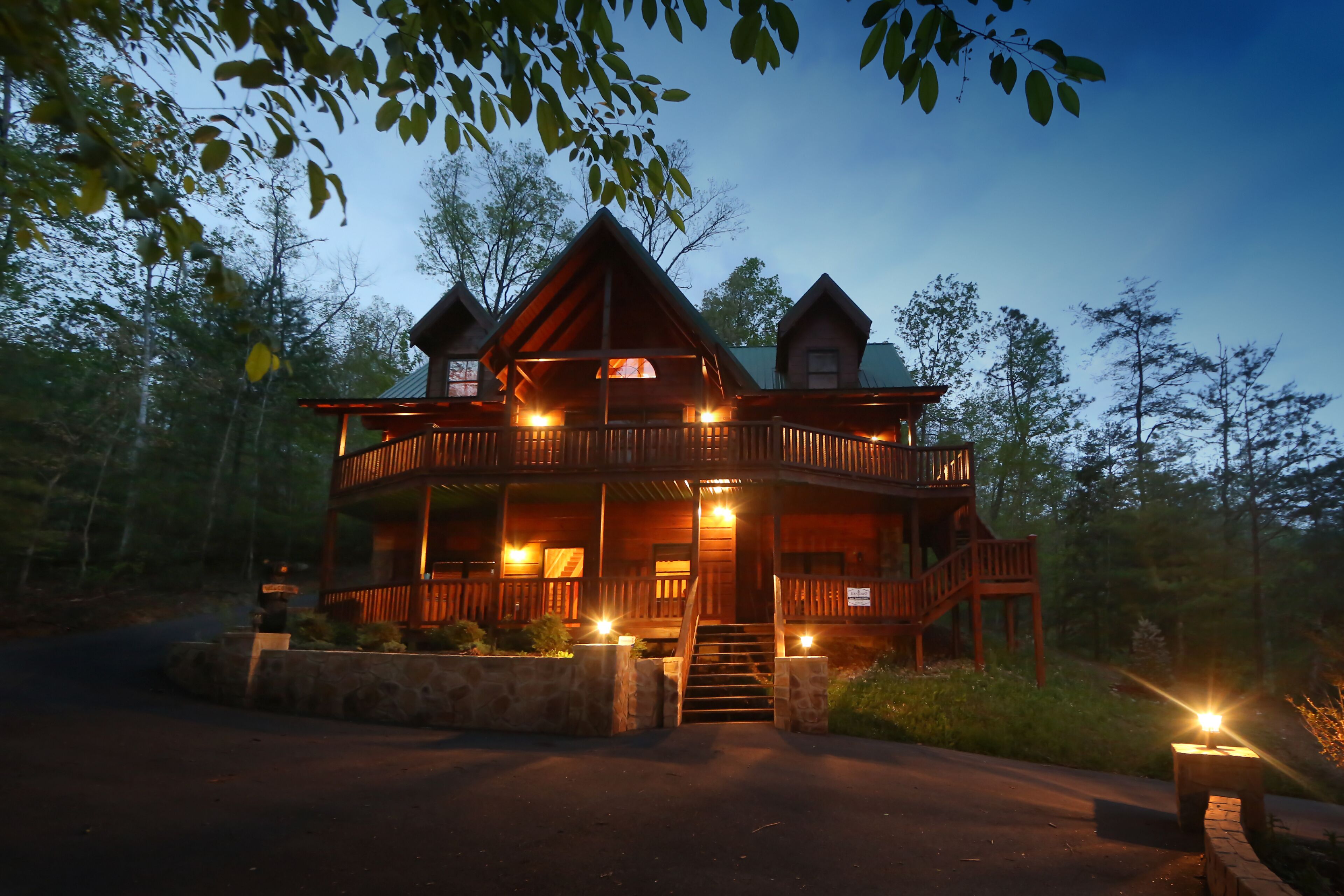 A two-story log cabin with a wrap-around porch and a second-story balcony, surrounded by trees.