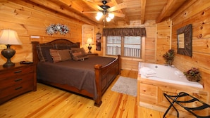Ein Schlafzimmer in einer Holzhütte mit einem Bett, Nachttisch, Lampe, Fenster mit Jalousien sowie einem Badezimmer mit Badewanne und Dusche.