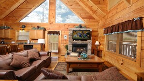 Ein gemütliches Blockhaus-Interieur mit Wohnzimmer, Kamin, Holzbalkendecke und großen Fenstern.