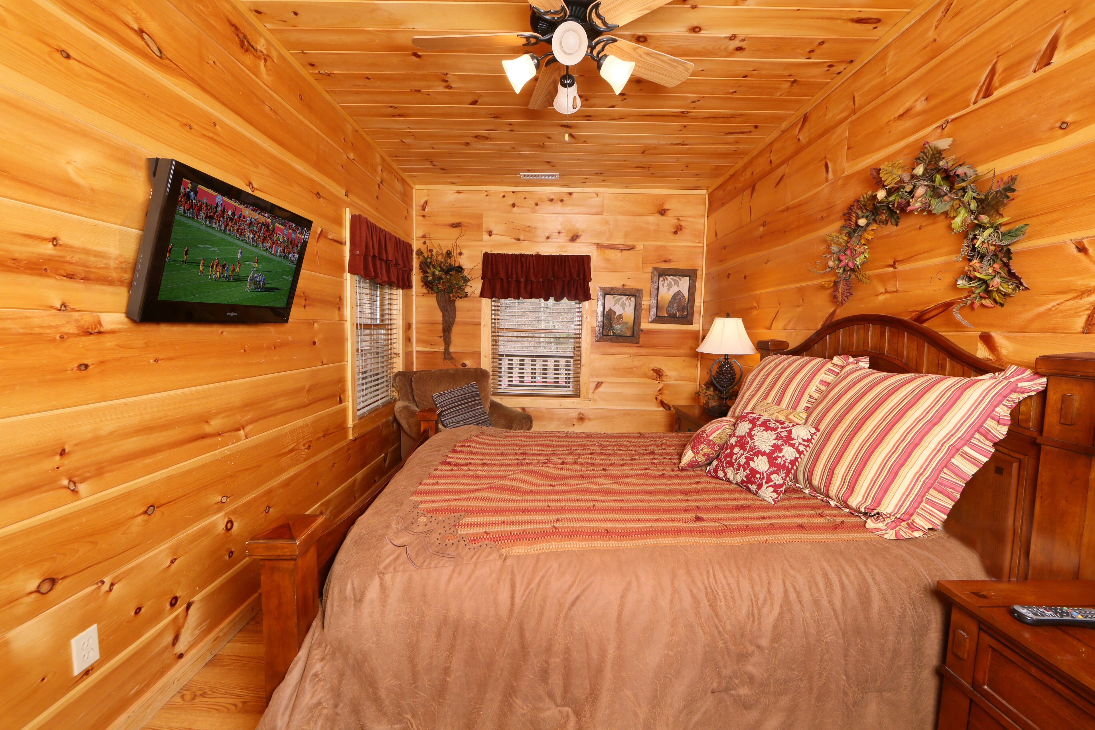 A cozy log cabin bedroom with a bed, a television, a ceiling fan, and a window with blinds.