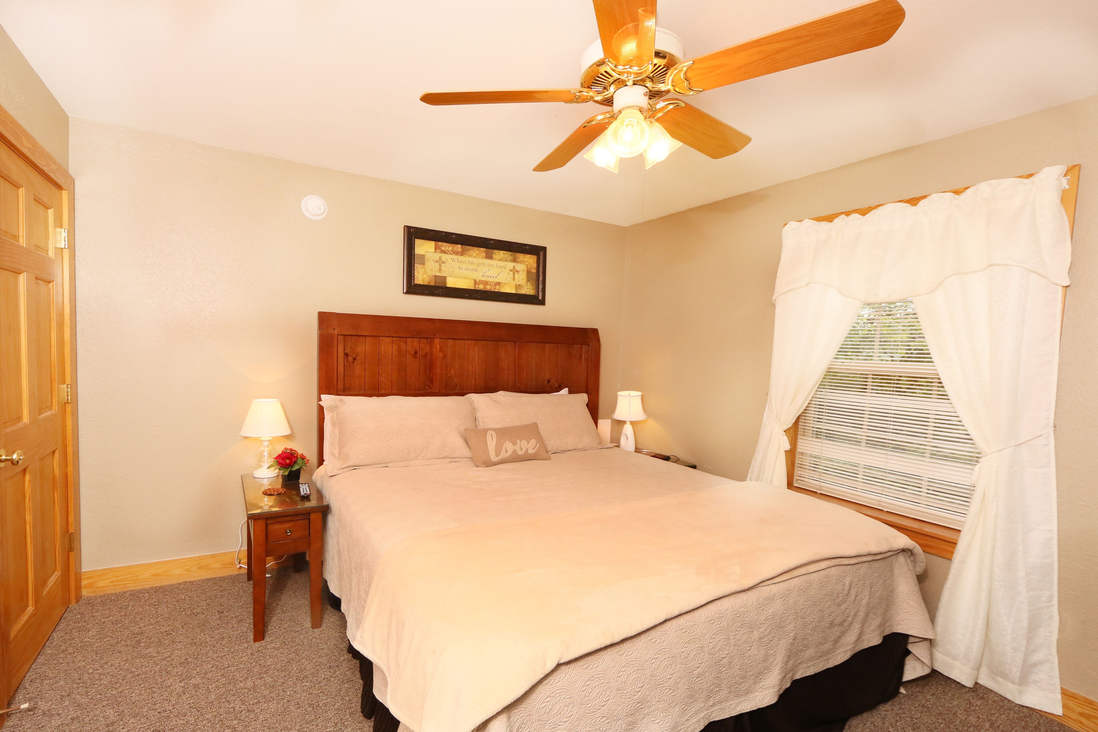 A bedroom with a wooden headboard, a ceiling fan, a bedside table with a lamp, a framed inspirational quote, and a window with white curtains.
