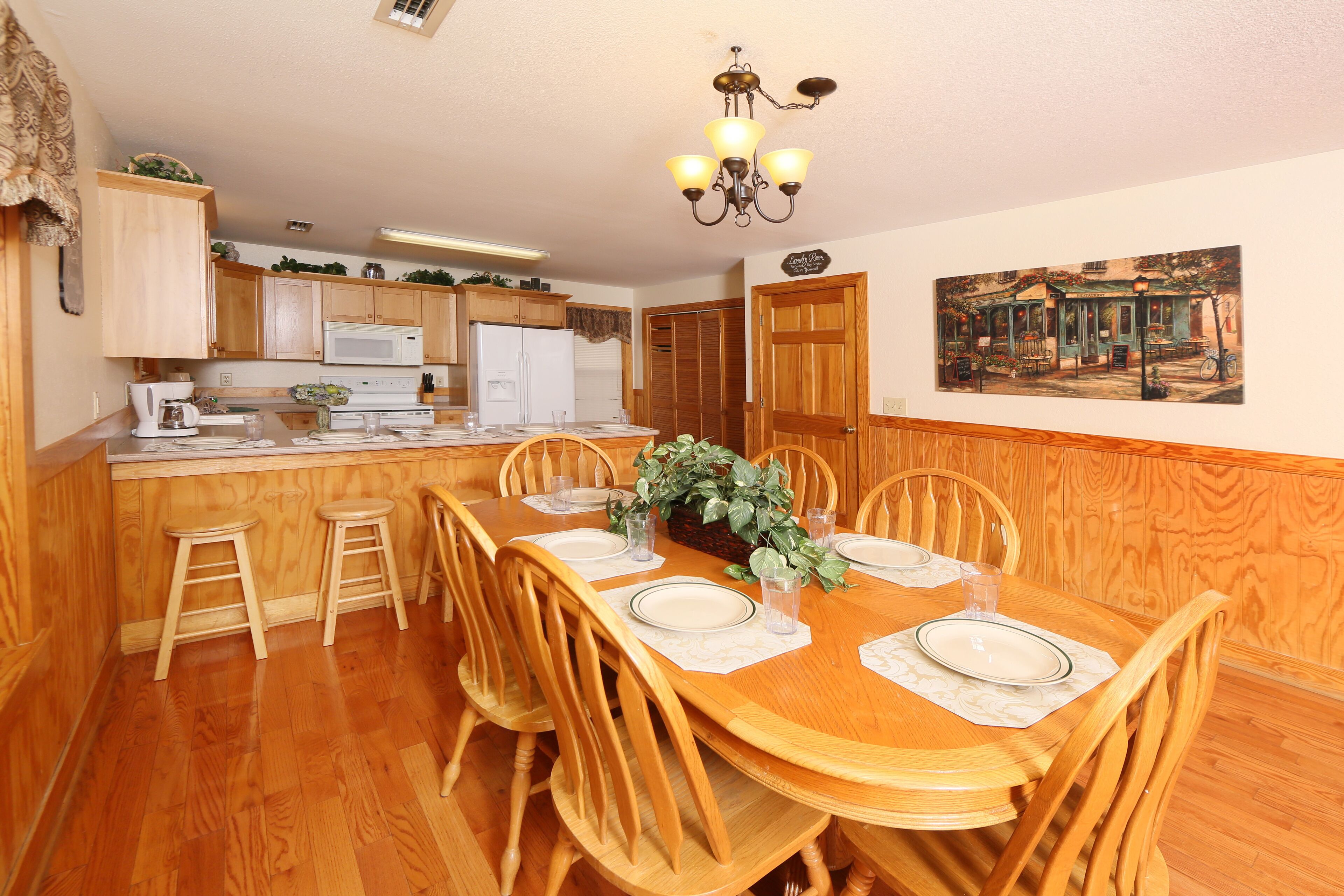 Un comedor con una mesa de madera dispuesta para seis personas, rodeada de sillas de madera. Un candelabro cuelga sobre la mesa, y hay un cuadro en la pared.