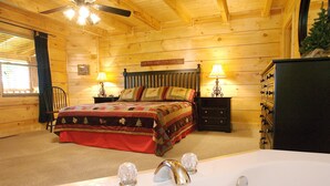 A wooden cabin bedroom with a bed, two bedside tables with lamps, a dresser, a ceiling fan, and a bathtub.