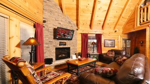 A cozy log cabin interior with a fireplace, leather sofa, wooden armchair, and a television mounted on the wall.