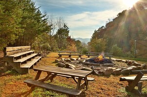 Um camping com mesas de piquenique, lareira e paisagem natural com árvores e vista para uma montanha ao longe.