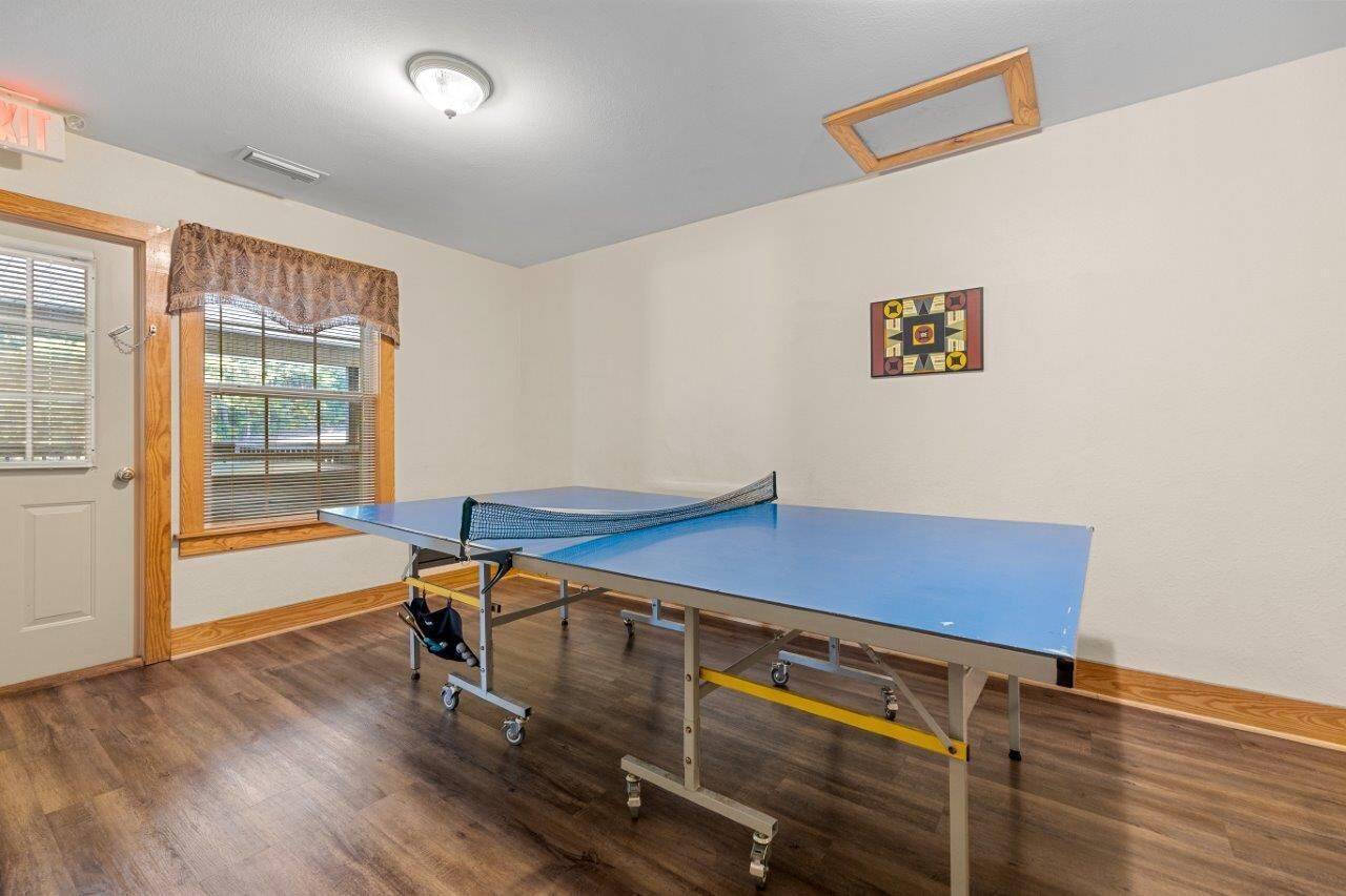 Una mesa de ping pong en una habitación con piso de madera, una ventana con persianas y una puerta.