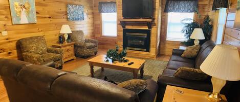 A cozy log cabin interior with a fireplace, leather sofas, and a flat-screen TV.
