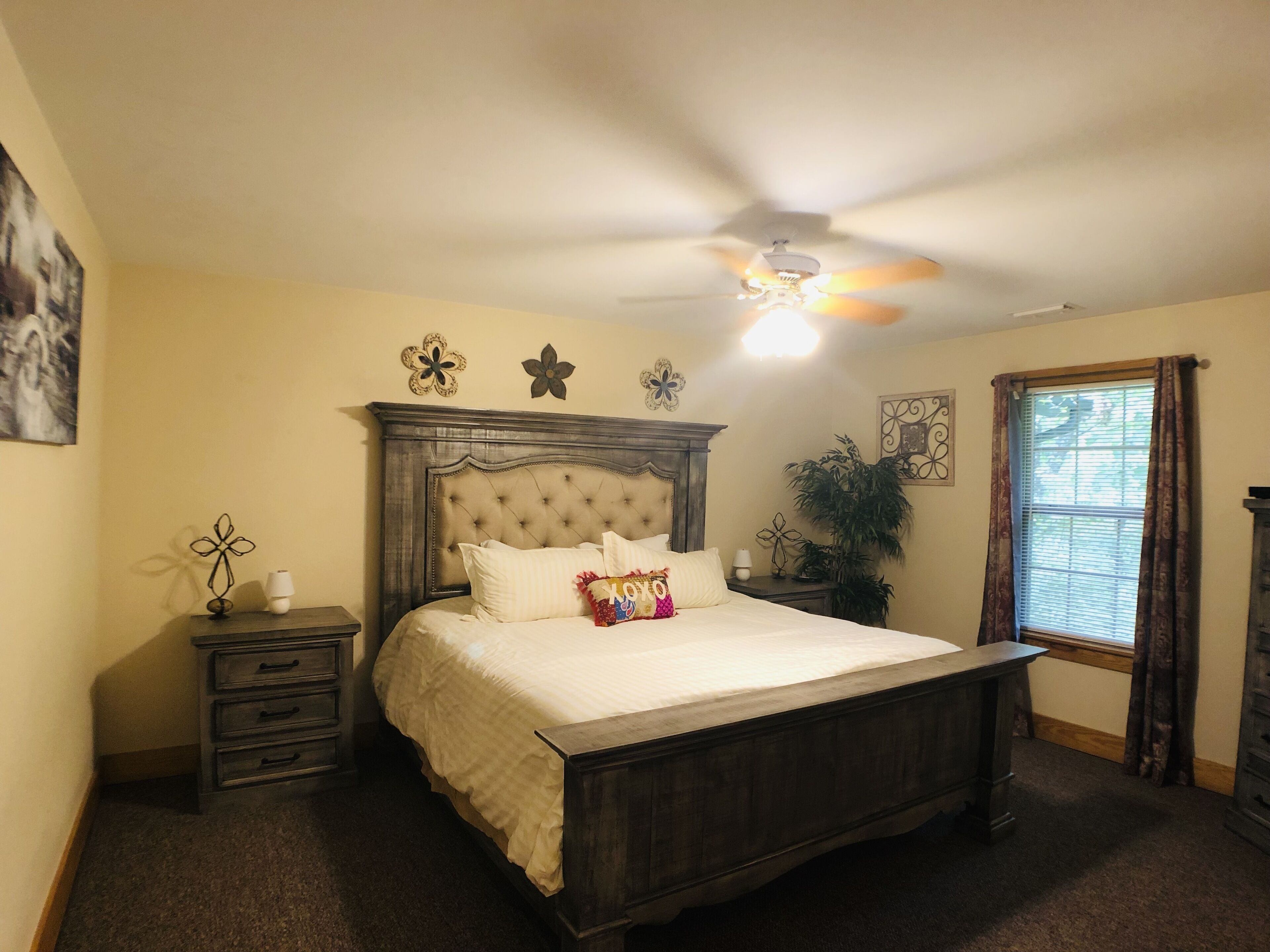 A bedroom with a tufted headboard, a bed with white linens, a bedside table with a lamp, a nightstand, a ceiling fan, and a window with curtains.