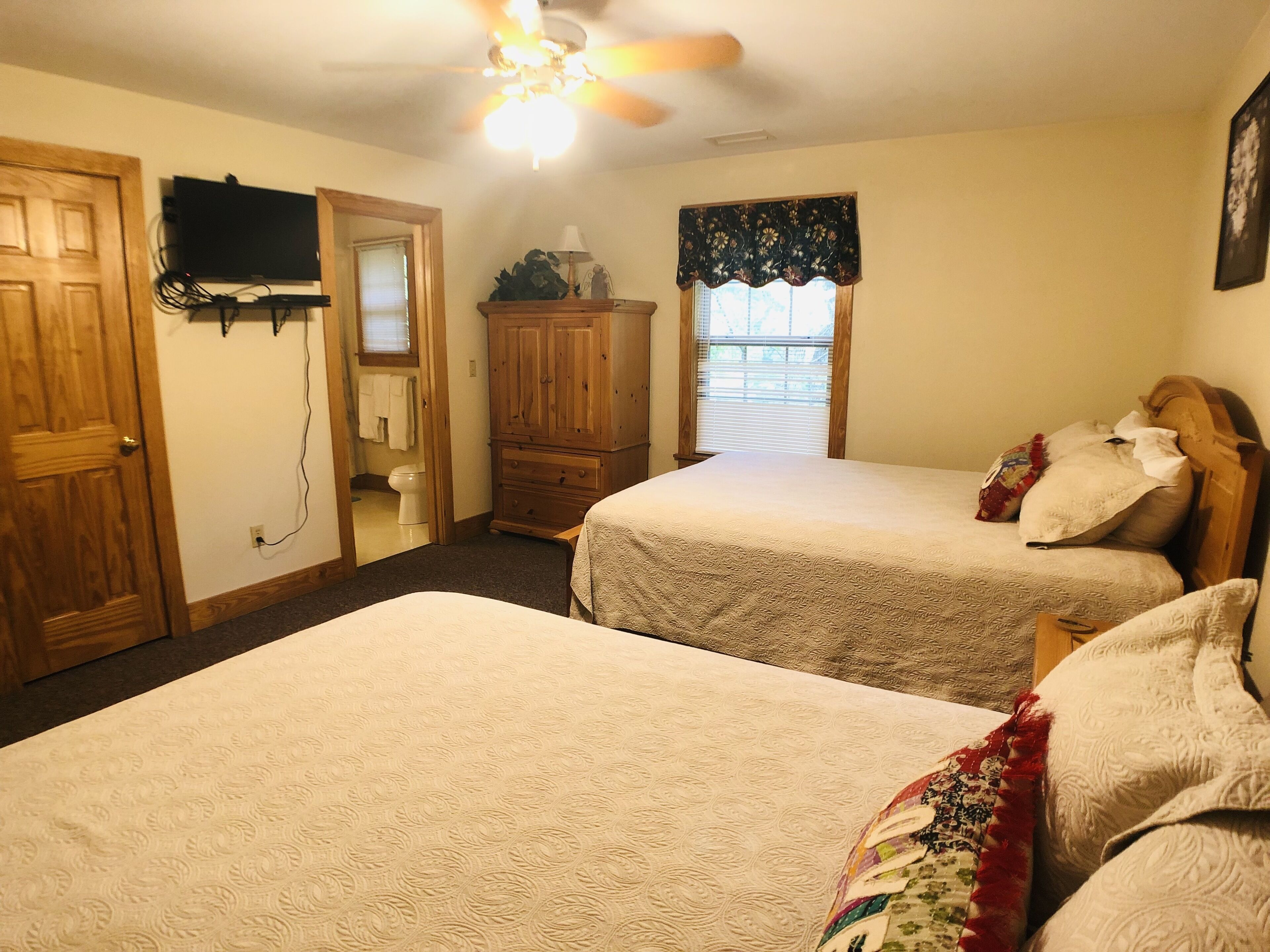 A bedroom with two beds, a wooden wardrobe, a television mounted on the wall, and a ceiling fan.
