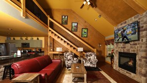 Ein geräumiges Wohnzimmer mit einem roten Ledersofa, zwei gemusterten Sessel, einem Holztisch und einem Kamin. Eine Treppe führt zu einer oberen Ebene, und ein Flachbildfernseher ist an der Wand befestigt.
