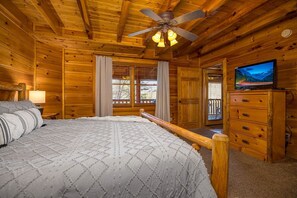 Una habitación tipo cabaña de madera con cama, ventilador de techo, televisión y ventana con cortinas.