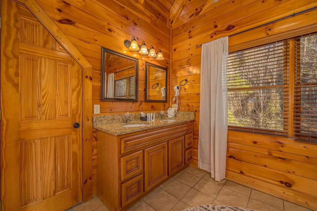 A wooden cabin bathroom with a vanity, mirrors, and a window with blinds.