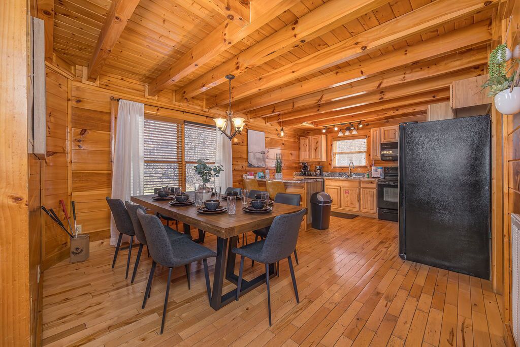 A wooden cabin interior with a dining area, kitchen, and a black refrigerator.