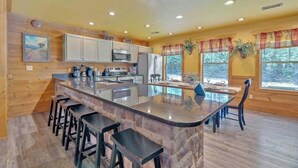 A kitchen with a large island, bar stools, wooden cabinets, and a view of the outdoors through large windows. - Splash Mountain Lodge (Gatlinburg)