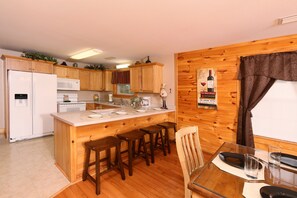 A kitchen with wooden cabinets, a refrigerator, microwave, and a dining area with a table and bar stools. - Pinnacle View 7B - Three Bedroom Chalet (Pigeon Forge)