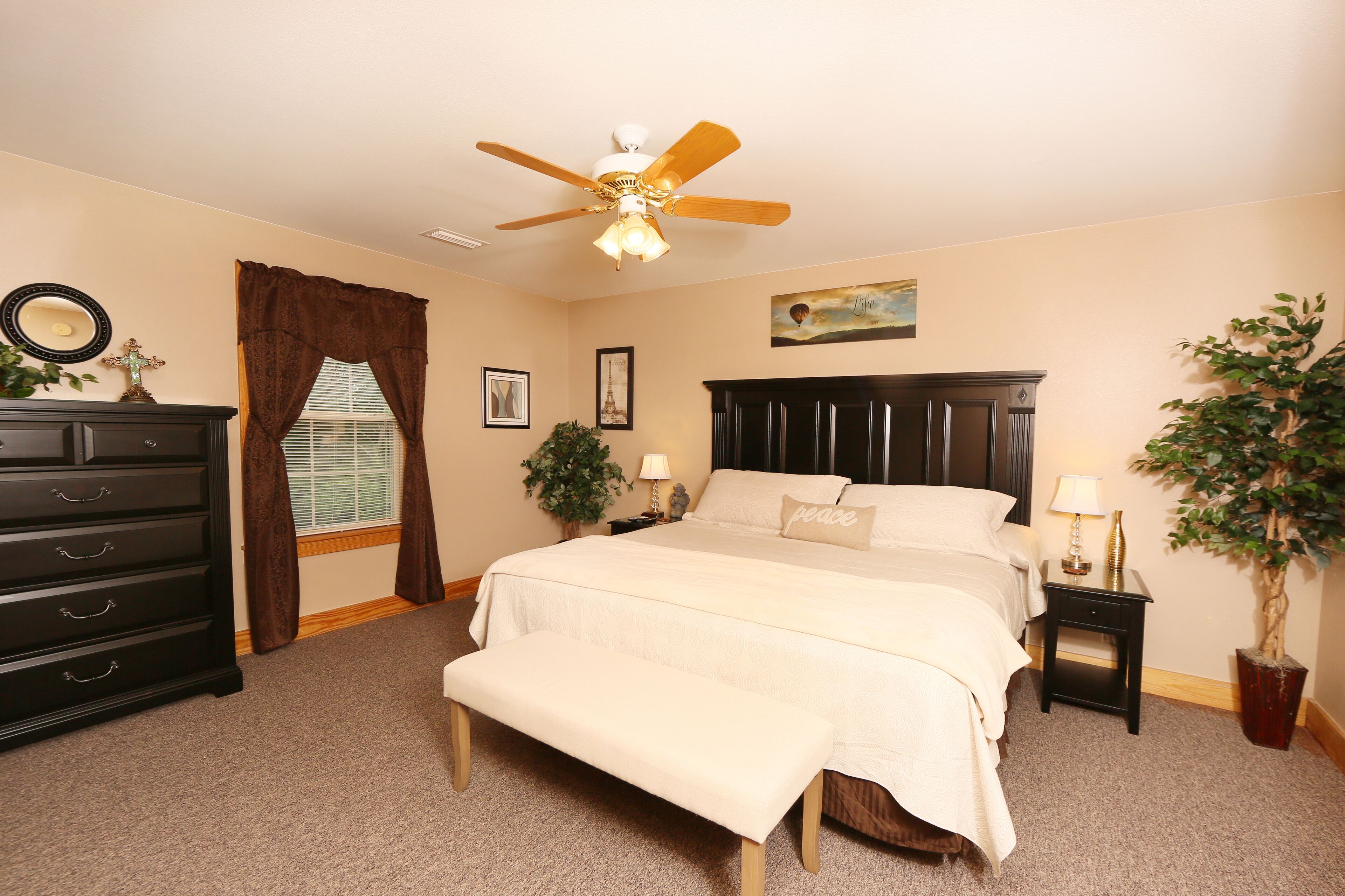 A bedroom with a bed, a dresser, a nightstand, a ceiling fan, and a potted plant.