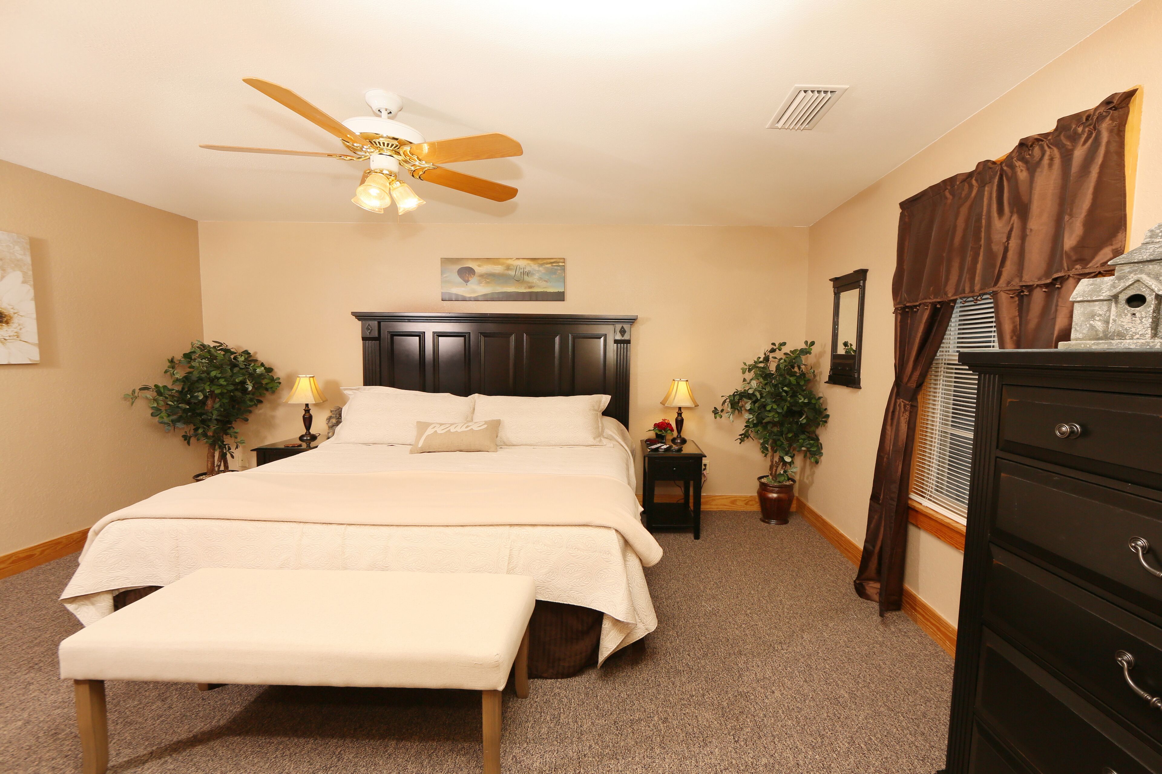 A bedroom with a bed, a bench, a ceiling fan, two nightstands with lamps, a dresser, and a window with curtains.