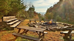 Un camping con mesas de picnic, un pozo de fuego y un paisaje natural con árboles y una vista distante de la montaña.