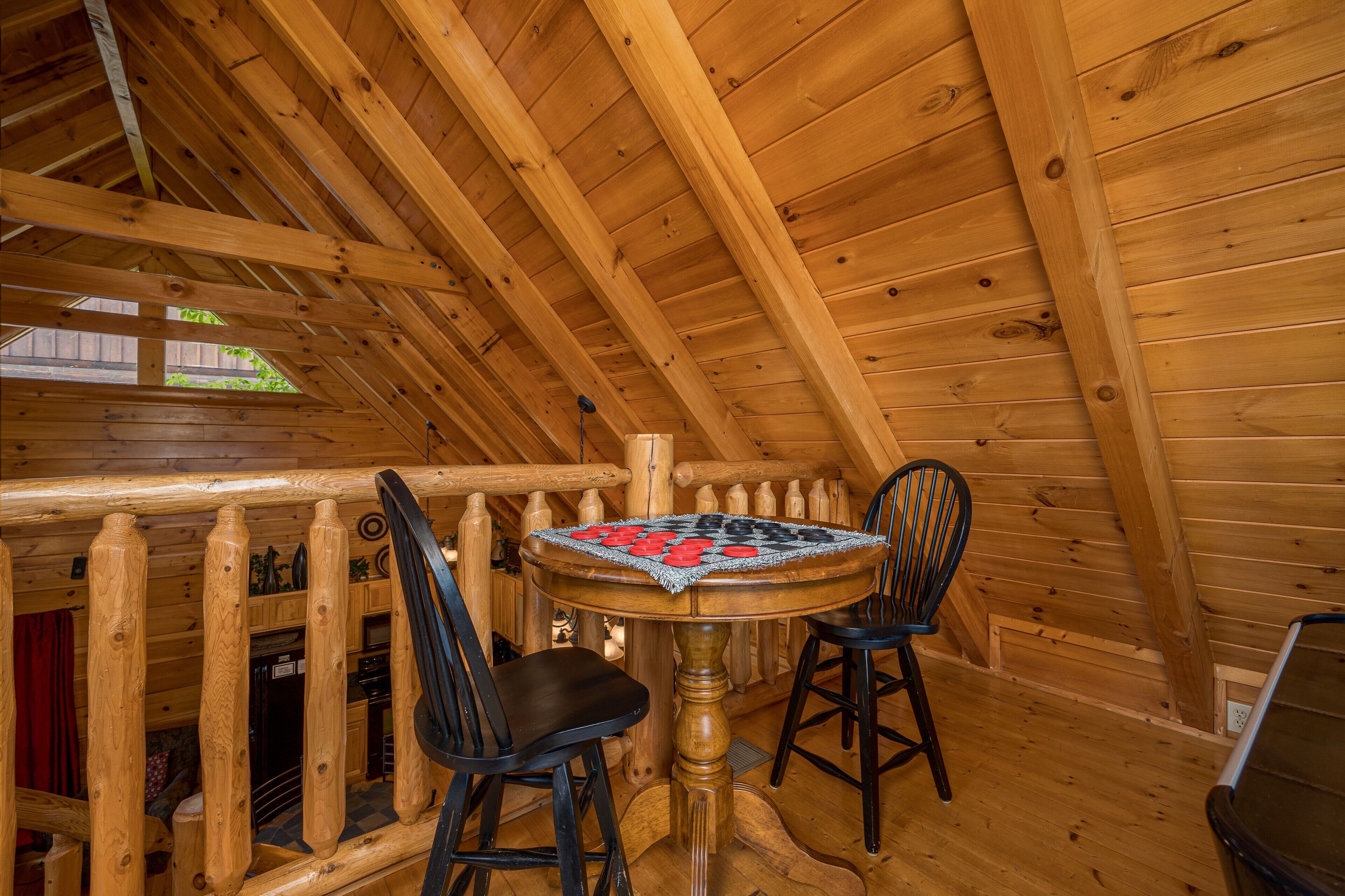 A wooden cabin interior with a dining table and chairs, a wooden staircase, and a view of greenery outside.