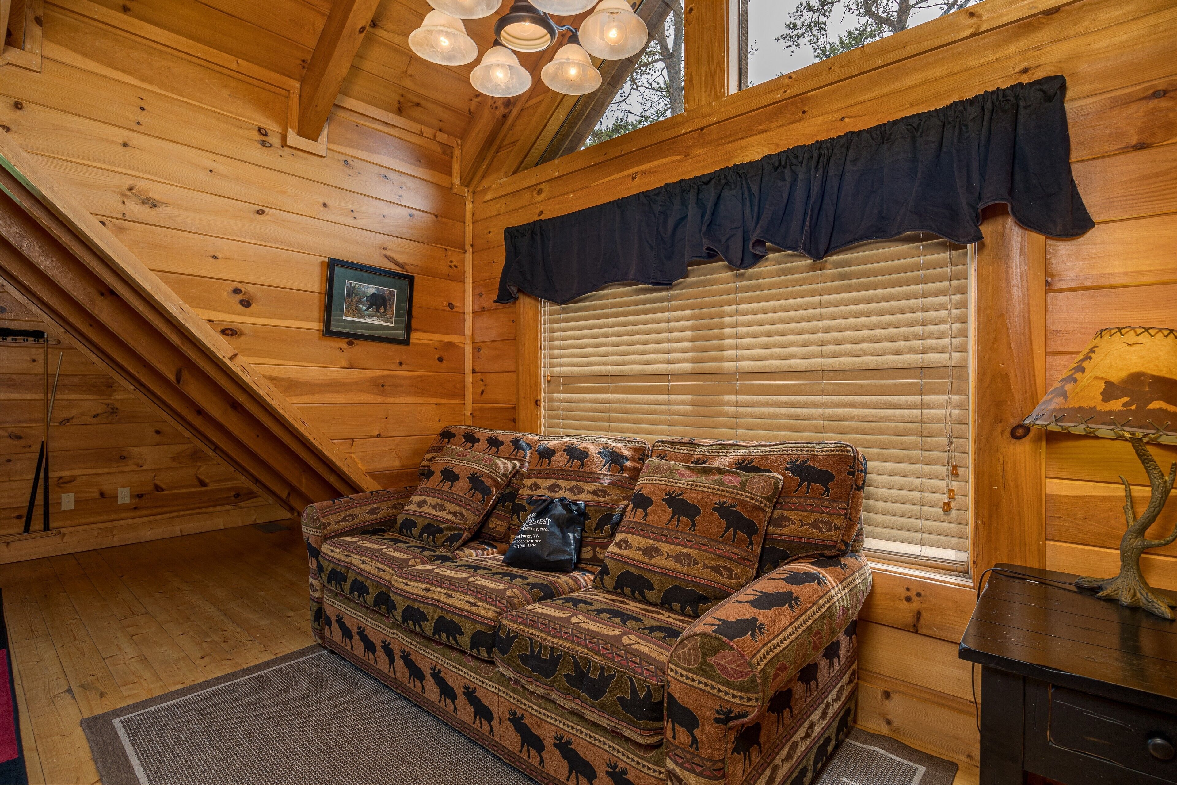 L’intérieur d’un chalet accueillant, avec un plafond en bois, une fenêtre équipée de stores, un tableau encadré au mur et un canapé à motifs.