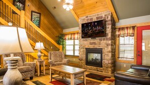 A cozy living room with a stone fireplace, a wooden staircase, and patterned furniture.