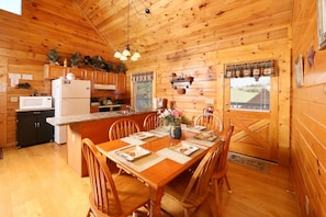Cozy cabin interior with a warm atmosphere, featuring a kitchen with wooden cabinetry and a dining area with a wooden table for four.
