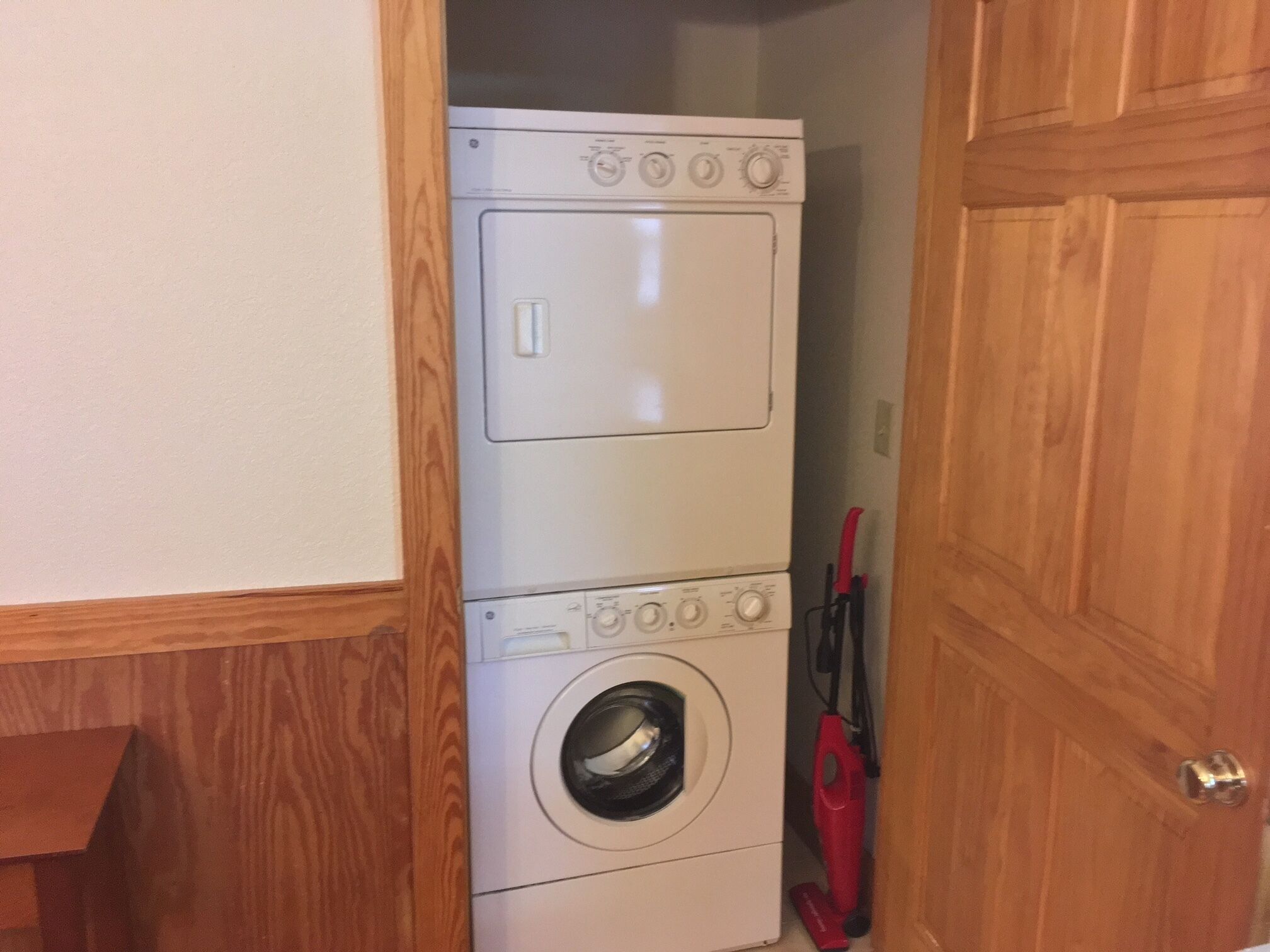 Une petite buanderie équipée d’un lave-linge et d’un sèche-linge superposés, de meubles en bois et d’un aspirateur rouge.