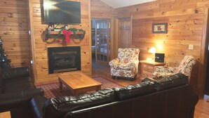 A cozy living room with a fireplace, leather sofa, armchairs, and a television mounted on the wall.