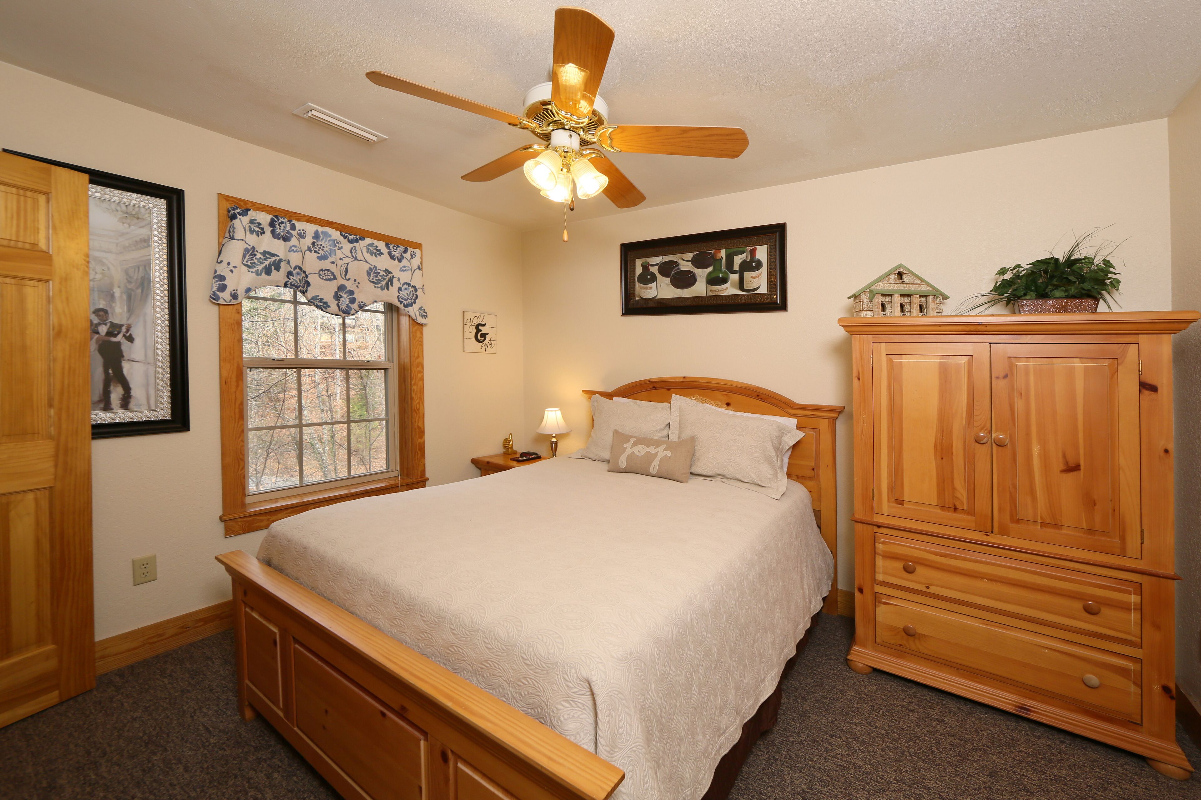 Une chambre à coucher comprenant un lit, une tête de lit en bois, une table de chevet, une lampe, un ventilateur de plafond, une fenêtre agrémentée d’une lambrequin à motifs floraux, des tableaux encadrés et une commode en bois.