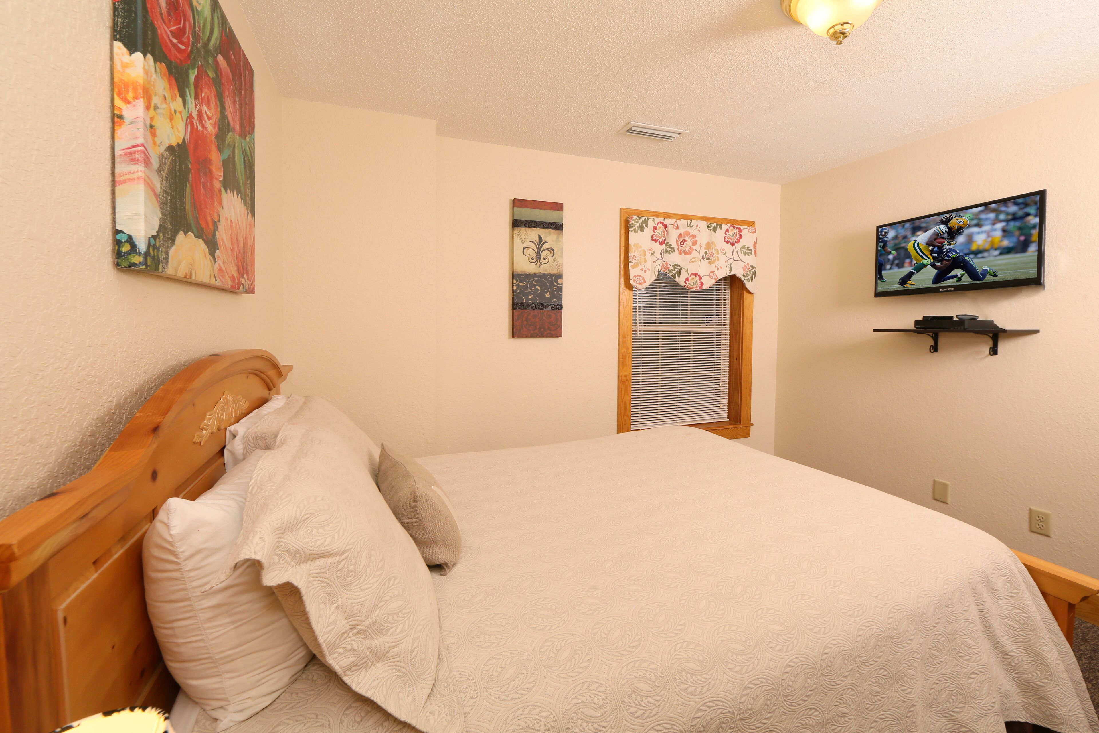 A bedroom with a wooden headboard, a bed with a white bedspread, a television mounted on the wall, and a window with a floral curtain.