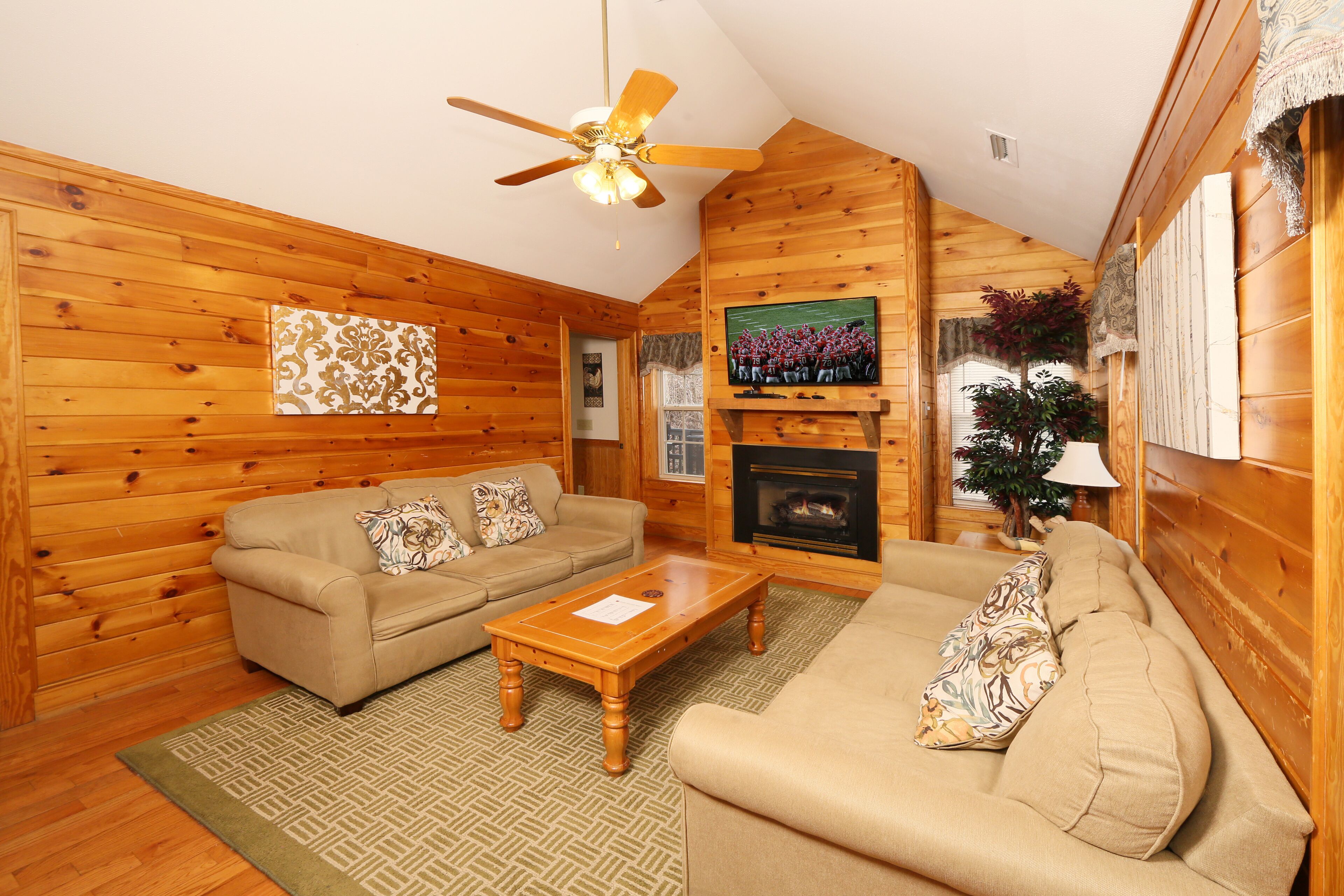 A cozy log cabin interior with a wooden floor, two beige sofas, a wooden coffee table, a ceiling fan, a fireplace, and a television mounted on the wall.