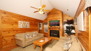 A cozy log cabin interior with a wooden floor, two beige sofas, a wooden coffee table, a ceiling fan, a fireplace, and a television mounted on the wall. - Ivy Falls 9 - Five Bedroom Chalet (Pigeon Forge)