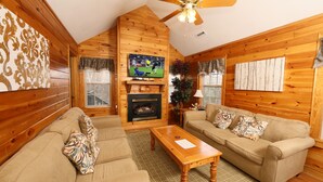 A cozy log cabin interior with a fireplace, wooden walls, a ceiling fan, and two sofas with patterned cushions. - Ivy Falls 9 - Five Bedroom Chalet (Pigeon Forge)
