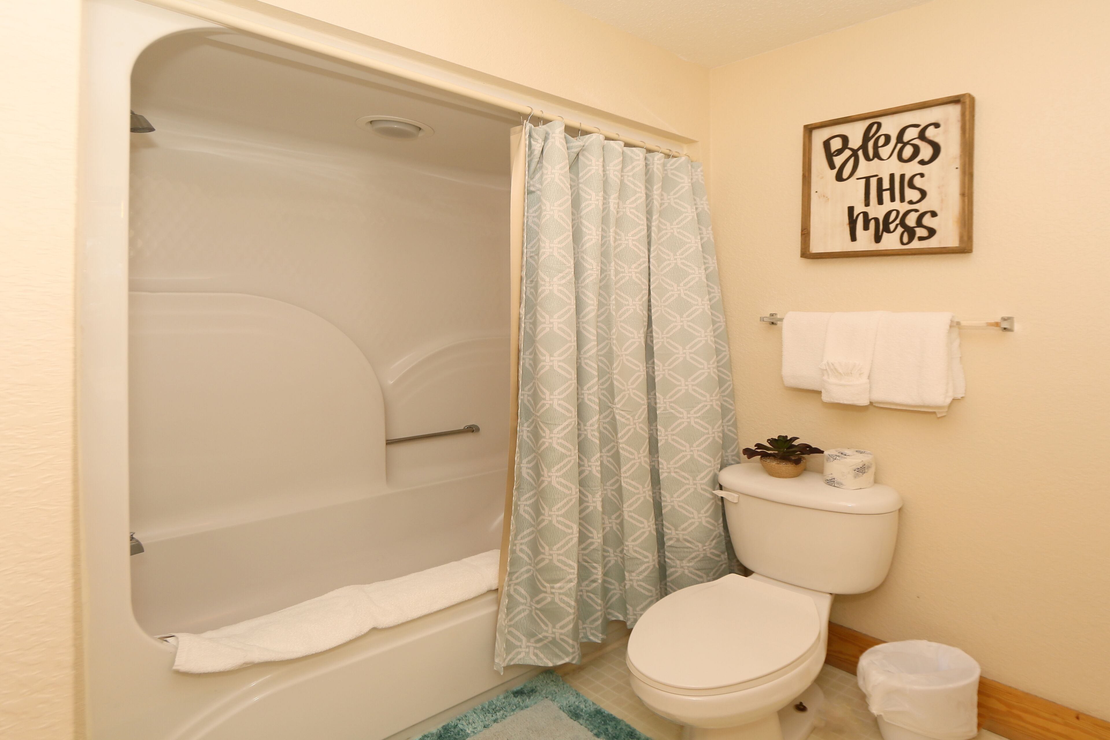 A bathroom with a bathtub, toilet, and a shower curtain. There is a sign on the wall that reads 'Bless this mess.'