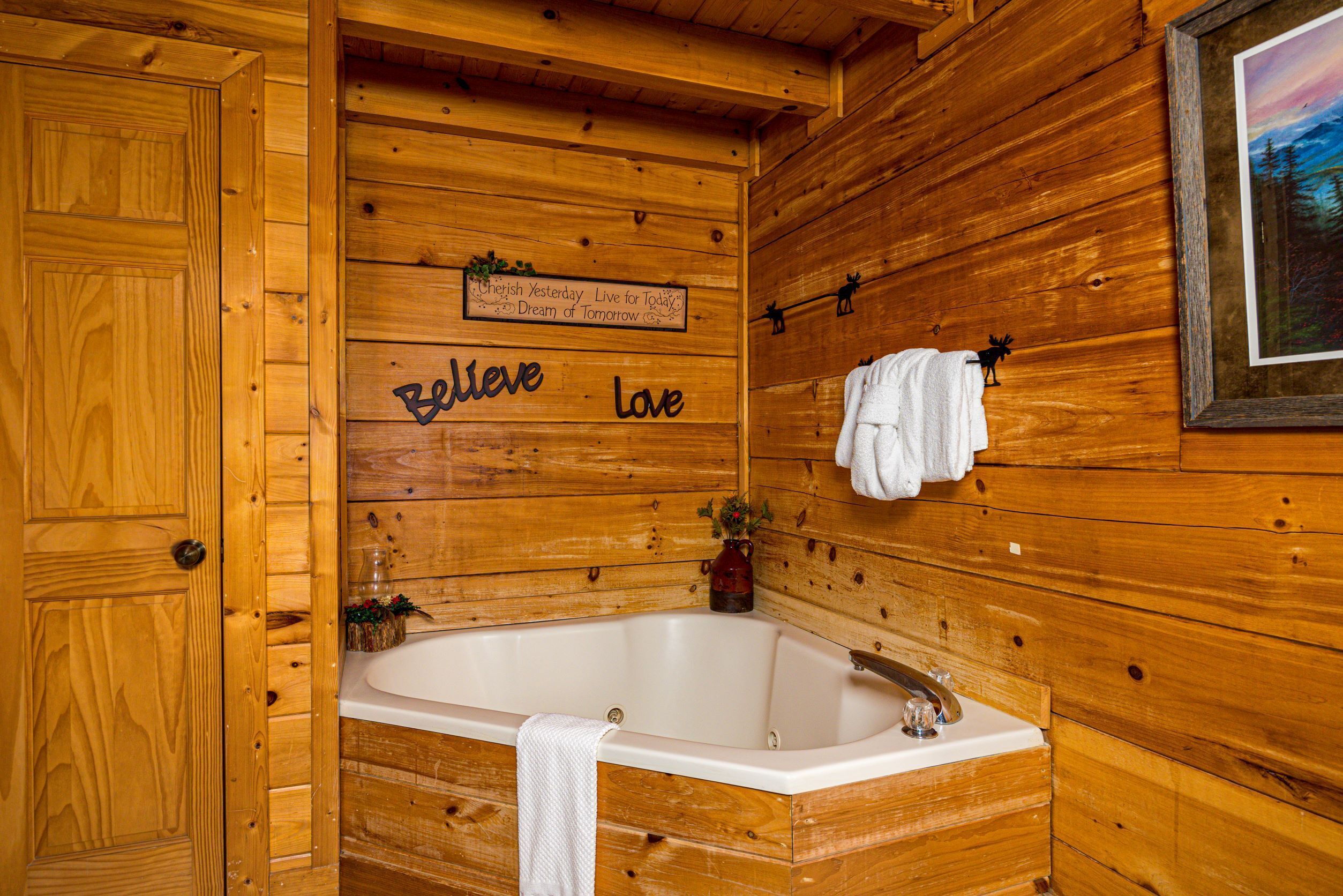 A wooden cabin interior with a wooden wall, a sign that says "Believe Love", a white bathtub, and a framed picture on the wall.