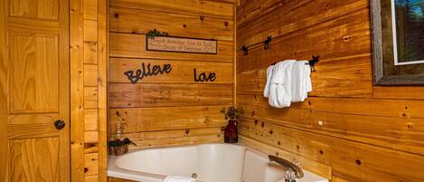 A wooden cabin interior with a wooden wall, a sign that says "Believe Love", a white bathtub, and a framed picture on the wall.