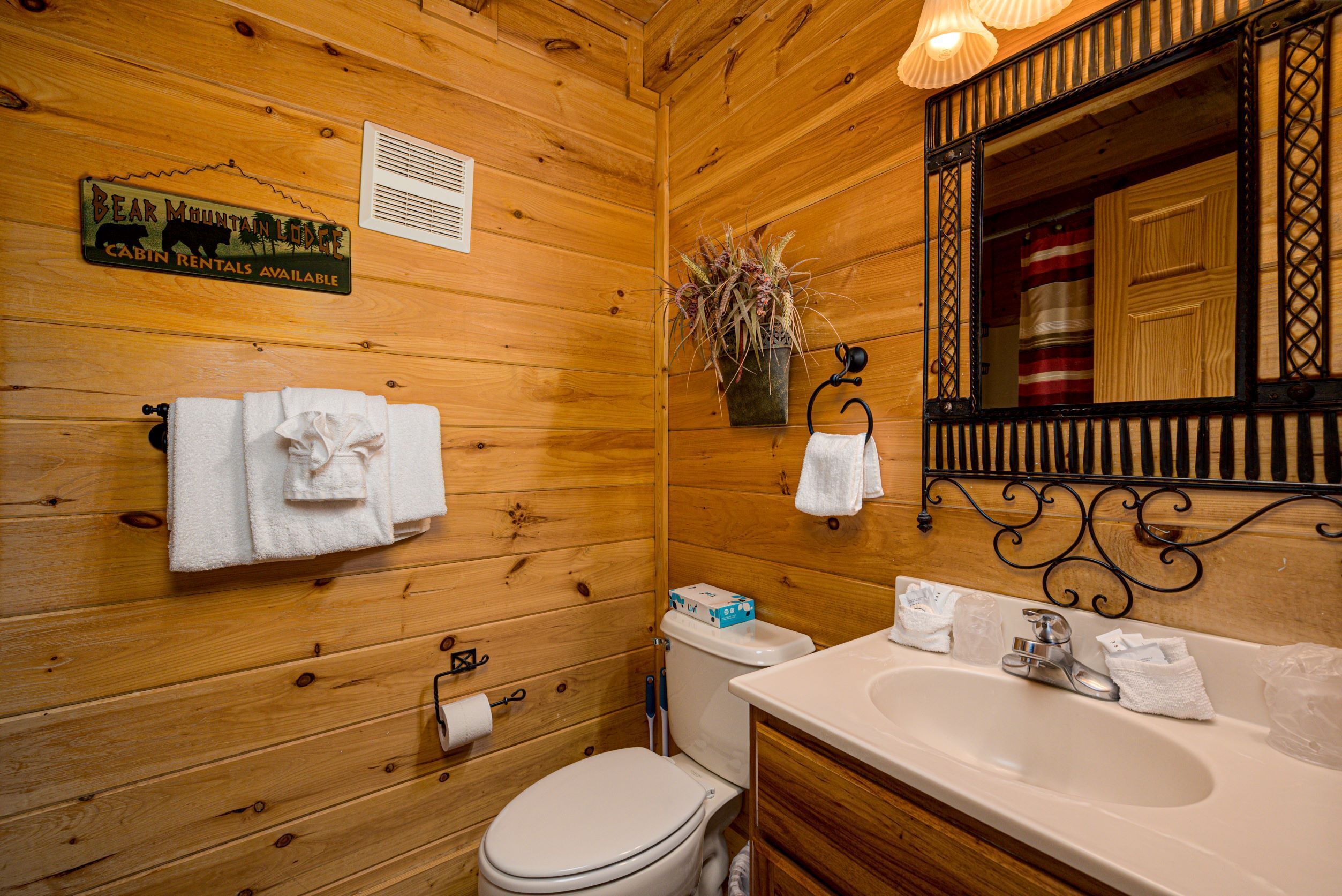 A wooden cabin bathroom with a toilet, sink, and a mirror. There are towels hanging on the wall and a sign indicating cabin rentals are available.