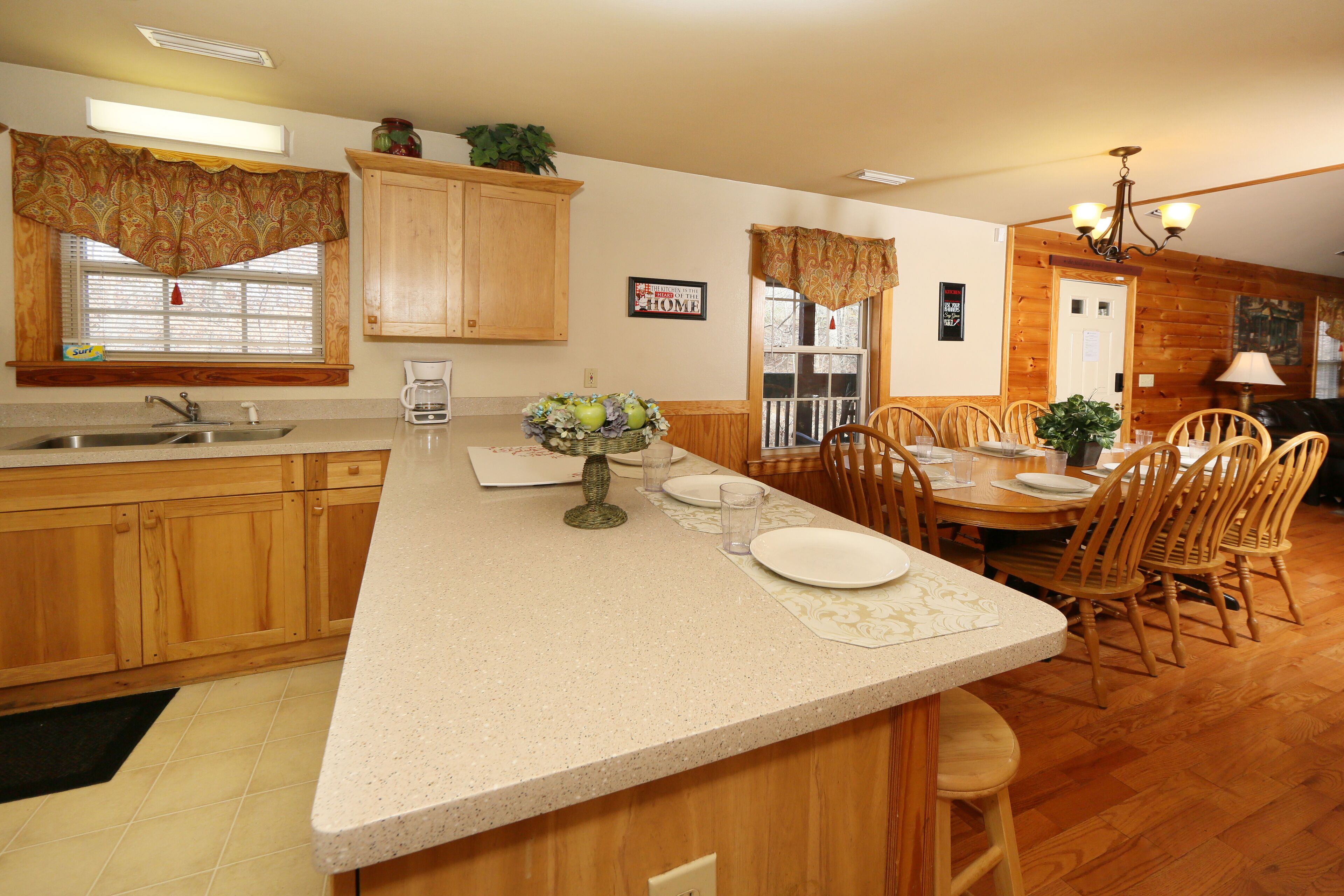 A kitchen with wooden cabinets, a large island with a vase of flowers, and a dining area with wooden chairs and a table set for four.